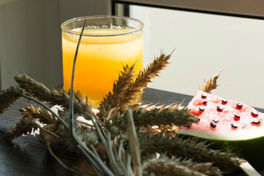 Freshly squeezed orange juice, sweet watermelon dessert and spikelets of ripe wheat on a wooden table in the sunlight...