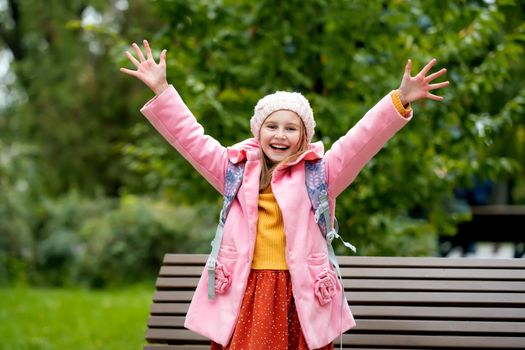 Pretty school girl kid with backpack looking at camera, holding hands up and smiling at autumn park. Beautiful female child portrait