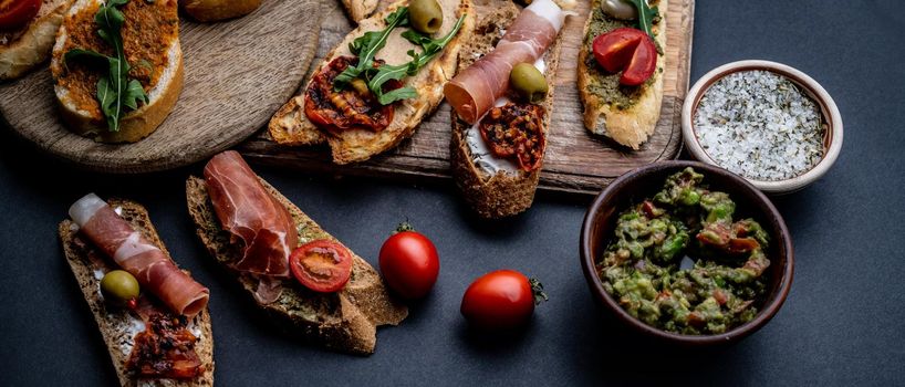 Set of bruschettas with jamon, olives, pesto, tomatoes, basil and mozzarella served on wooden board with guacamole and salt with herbs. Traditional mediterranean antipasti food composition