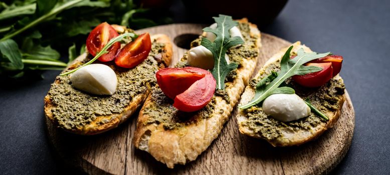 Bruschettas with pesto, tomatoes, mozzarella cheese and basil on wooden board served with guacamole, arugula and salt with herb. Italian tosted bread with vegetables and traditional sauce