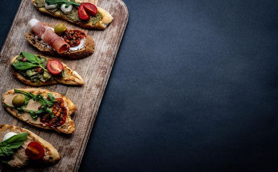 Bruschettas with jamon, olives, pesto, grilled cherry tomatoes served on wooden board with arugula on background with copy space. Mediterranean toasted bread with meat, cheese and vegetables