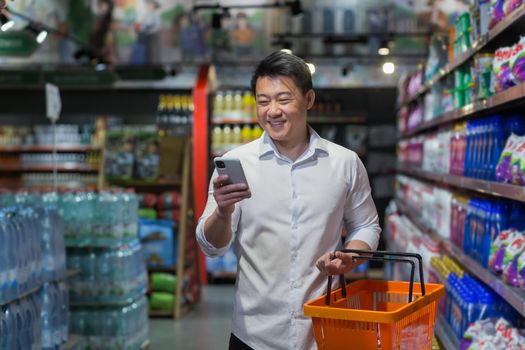 Experienced mature Asian shopper in supermarket, man smiling and reading message on smartphone, makes purchases and chooses products from various shelves