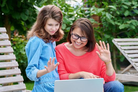 Mom and teenage daughter using laptop for chat conference video call. Parent and child relaxing together in backyard outdoor on lounger, laughing, waving hand at screen. Family technology lifestyle