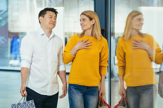 multiracial couple asian man and caucasian woman walking together in mall after shopping with colored bags in hands. walking past shop windows with clothes cheerful family hugging smile.