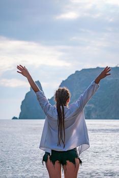 The girl stands on the shore and looks at the sea. Her hands are raised up. She wears a white shirt and her hair is in a braid