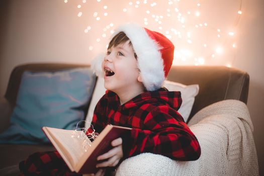 A boy in a Santa hat is reading a book on a lifestyle sofa. Reading books. New Year's mood. Garland on the wall