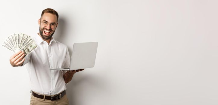 Business and e-commerce. Happy successful businessman bragging with money, working on laptop online, standing over white background.