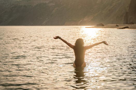 The girl stands in the sea and looks ahead at the sunset. Her hands are raised up. She wears a black swimsuit and her hair is down