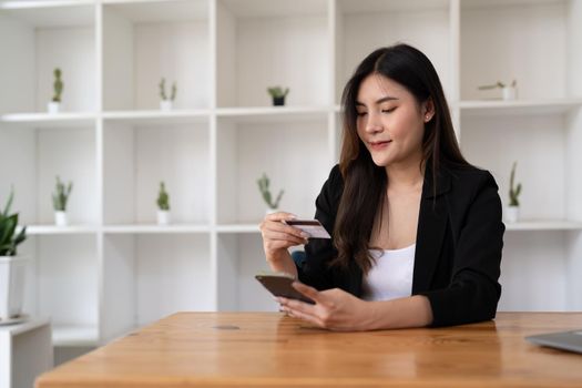 Young Asian woman using credit card and mobile phone for online shopping in coffee shop or coworking space, coffee cup, technology money wallet and online payment concept, credit card mockup.