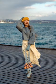 Outdoors fashion portrait of a beautiful middle aged woman walking on the beach. Marine background. Dressed in a stylish warm blue sweater, yellow skirt and beret