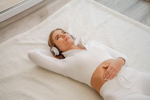 Top view portrait of relaxed woman listening to music with headphones lying on carpet at home. She is dressed in a White tracksuit