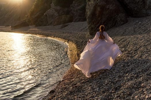 A mysterious female silhouette with long braids stands on the sea beach with mountain views, Sunset rays shine on a woman. Throws up a long white dress, a divine sunset