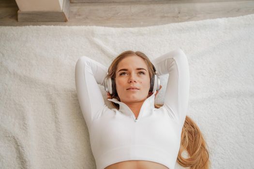 Top view portrait of relaxed woman listening to music with headphones lying on carpet at home. She is dressed in a White tracksuit