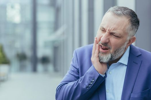 Toothache. Close-up photo. Senior man, businessman, office worker standing on the street near work, holding his cheek, feeling unbearable toothache, grimacing, needs help.
