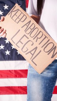 Young woman protester holds cardboard with Keep Abortion Legal sign against USA flag on background. Girl protesting against anti-abortion laws. Feminist power. Equal opportunity Womens rights reedom