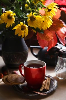 Red cup of tea on metal plate with a bouquet of yellow autumn flowers and colored leaves on white table, early autumn morning concept, selective focus.