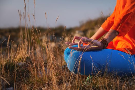 Close-up of a woman and female hands sitting in a lotus position, yoga outdoors in the grass at sunset against the backdrop of a mountain range and clouds, meditation.