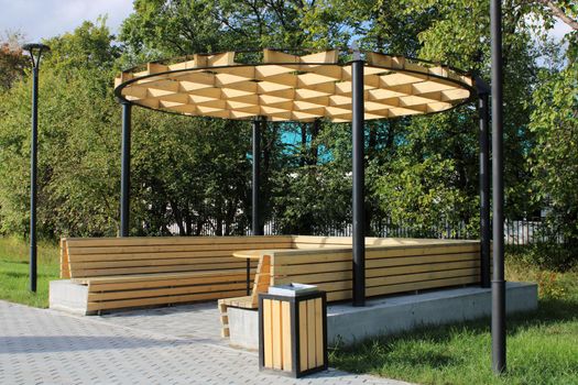 A light wooden gazebo with a bench, a table and an unusual hutch in a city park. Urban architecture.