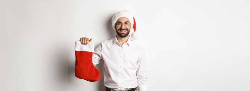 Merry christmas, holidays concept. Happy adult man receive gifts in xmas sock, looking excited, wearing santa hat, white background.
