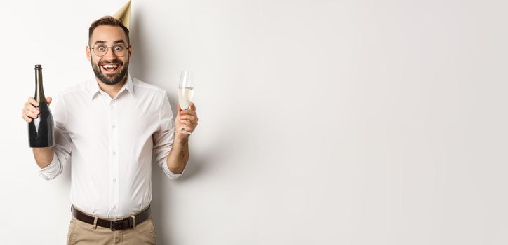 Celebration and holidays. Excited man enjoying birthday party, wearing b-day hat and drinking champagne, standing over white background.