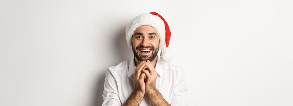 Party, winter holidays and celebration concept. Excited and hopeful man in santa hat looking at christmas gift with amazement, white background.