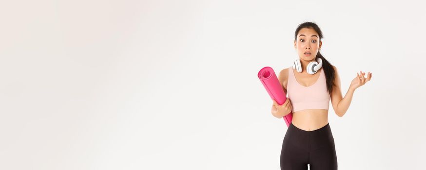Sport, wellbeing and active lifestyle concept. Confused and shocked asian fitness girl, cant understand why gym closed, holding rubber mat and looking surprised at camera, white background.