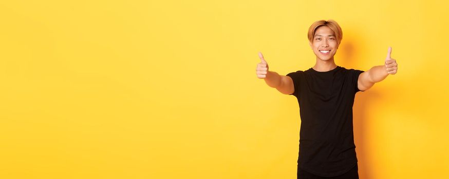 Portrait of assured handsome asian man showing thumbs-up in approval, like idea, standing yellow background.