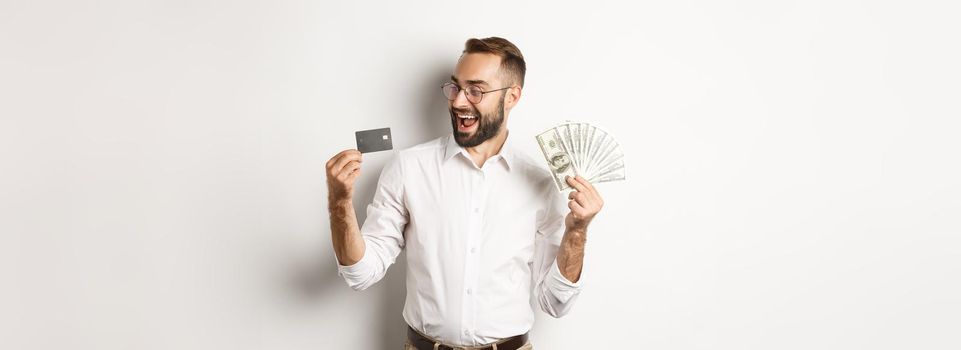 Excited businessman holding money and looking at credit card, standing over white background.