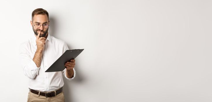 Thoughtful employee looking at clipboard, having doubts, standing over white background.