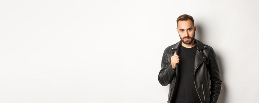 Handsome bearded man expressing confidence, touching his leather jacket and looking self-assured, white background.