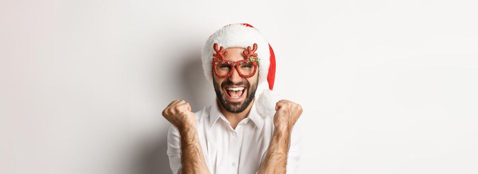 Close-up of man celebrating christmas or new year, wearing xmas party glasses and santa hat, rejoicing and shouting of joy, standing over white background.