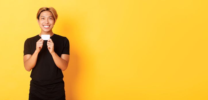 Excited happy asian guy smiling and showing credit card, standing yellow background.