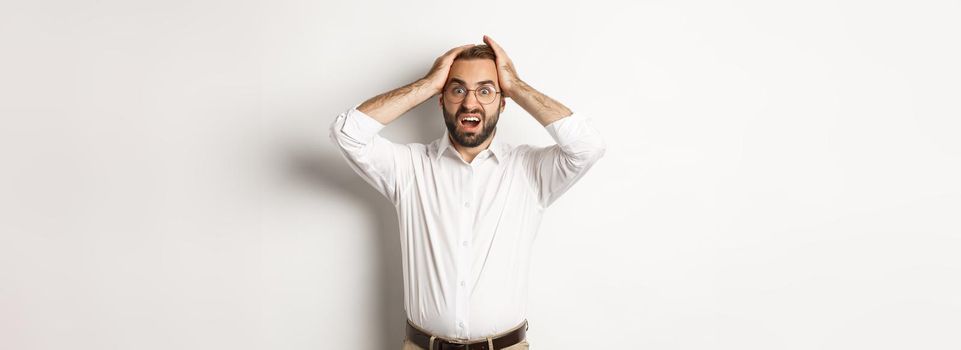 Frustrated businessman holding hands on head, looking shocked and anxious, standing over white background.