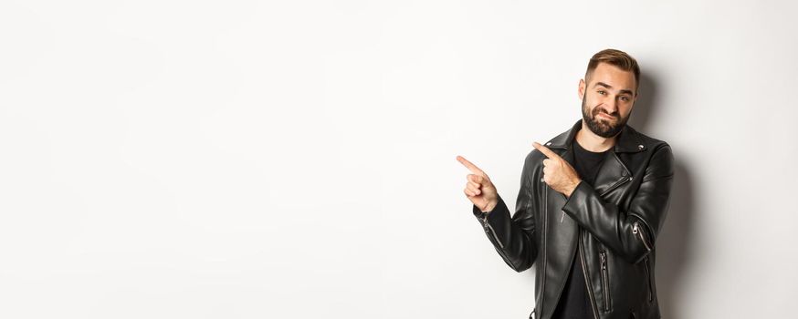 Skeptical and disapleased man in leather jacket, pointing fingers at upper left corner, showing bad promo offer, standing over white background reluctant.