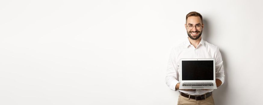 Handsome businessman in glasses, showing laptop screen and smiling happy, standing over white background.
