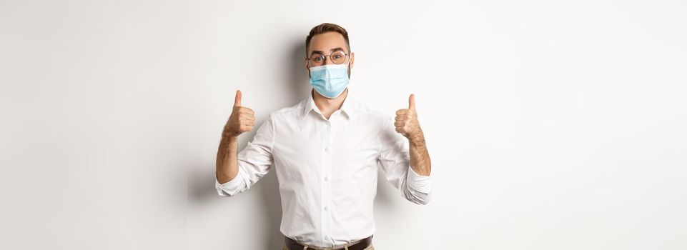 Covid-19, social distancing and quarantine concept. Satisfied male manager showing thumbs up, recommending to wear face mask, standing over white background.