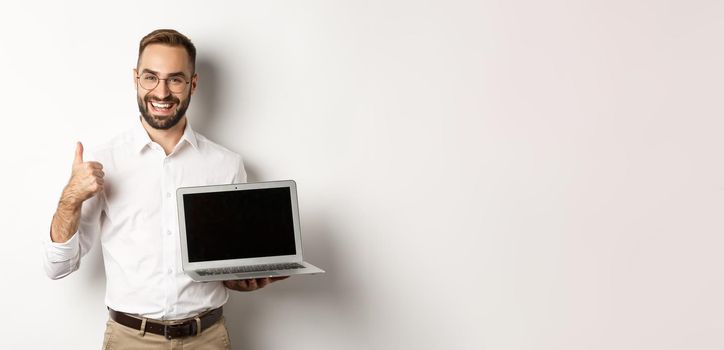 Successful business man showing laptop screen, make thumb up in approval, praise something good, standing over white background.