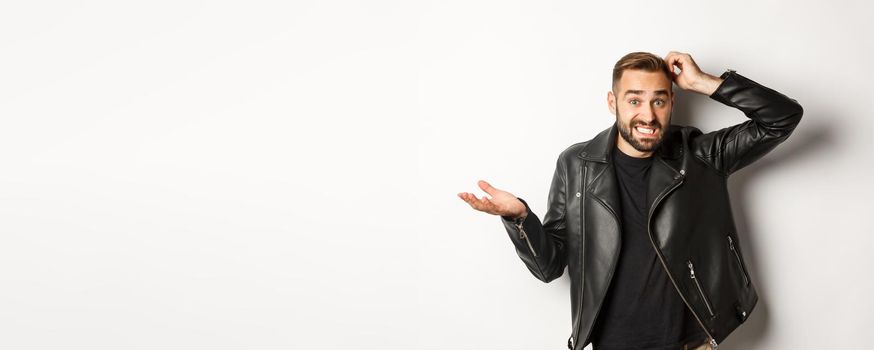 Confused bearded guy in cool leather jacket shrugging, scratch head puzzled and clueless, standing over white background.