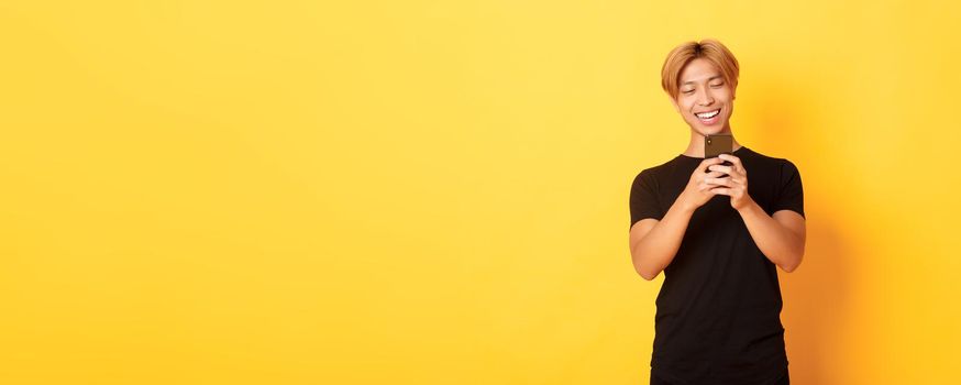 Portrait of handsome stylish asian guy with blond hair, using mobile phone and smiling, messaging in smartphone app, yellow background.