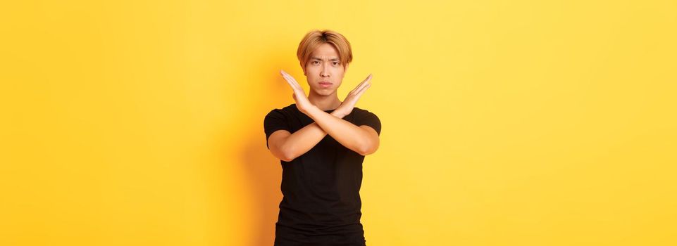 Portrait of disappointed frowning asian man, making cross gesture to stop something bad, standing yellow background.