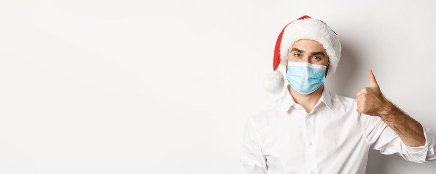 Concept of covid-19, social distancing and winter holidays. Satisfied man in face mask and santa hat showing thumb up, celebrating christmas with preventive measures, white background.