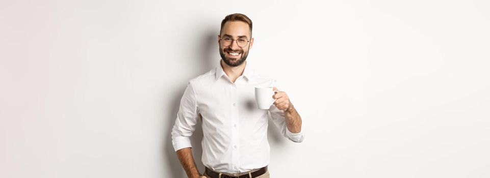 Handsome businessman drinking coffee and smiling, standing against white background.