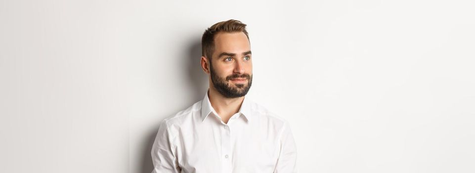 Close-up of confident businessman in white shirt, looking left and smiling satisfied, standing over white background.