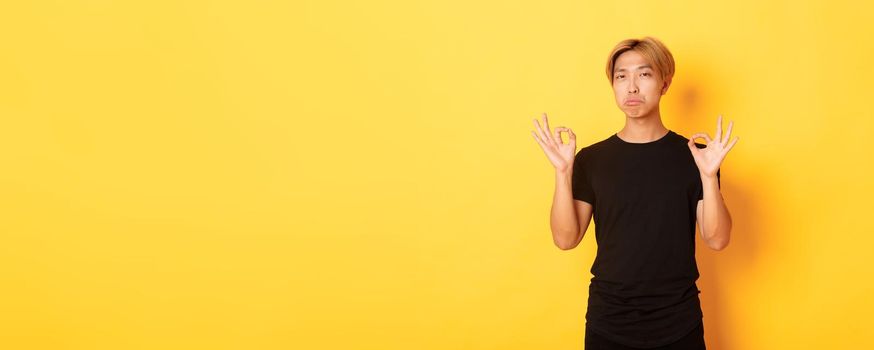 Impressed asian stylish guy with blond hair, showing okay gesture and praise something good, standing yellow background.