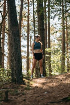 Portrait of young woman runner dressed in sportswear standing on trail in green forest, ready to run while preparing for marathon.