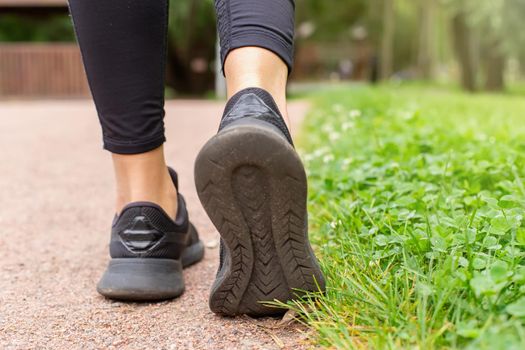 a womans legs, in black sneakers and black leggings, walking along a path made of small red stone, next to a green lawn with grass. Close up