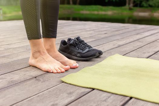 Close-up of bare feet on a wooden floor near a green sports mat, there are black sneakers nearby, in summer, near a pond in the park
