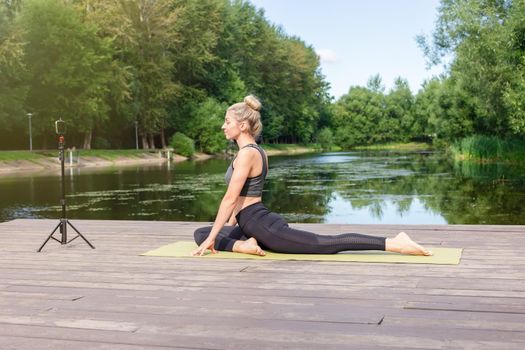 A slender beautiful woman in a gray top and leggings, on a wooden platform by a pond in the park in summer, does yoga, sits on a green sports mat, in front of her smartphone on a tripod.