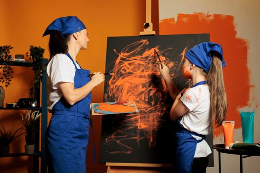 Woman and child creating masterpiece with orange color aquarelle paint and dye palette with brush. Little girl learning painting skills on canvas using watercolor design and artwork inspiration.