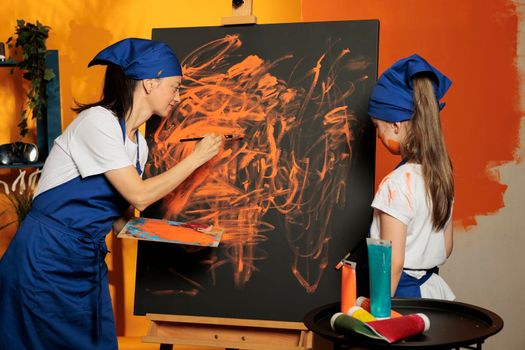 Woman teaching kid to paint canvas artwork with watercolor aquarelle and paintbrush, having creative skills. Painting artistic masterpiece design with watercolor dye and tools, art practice.